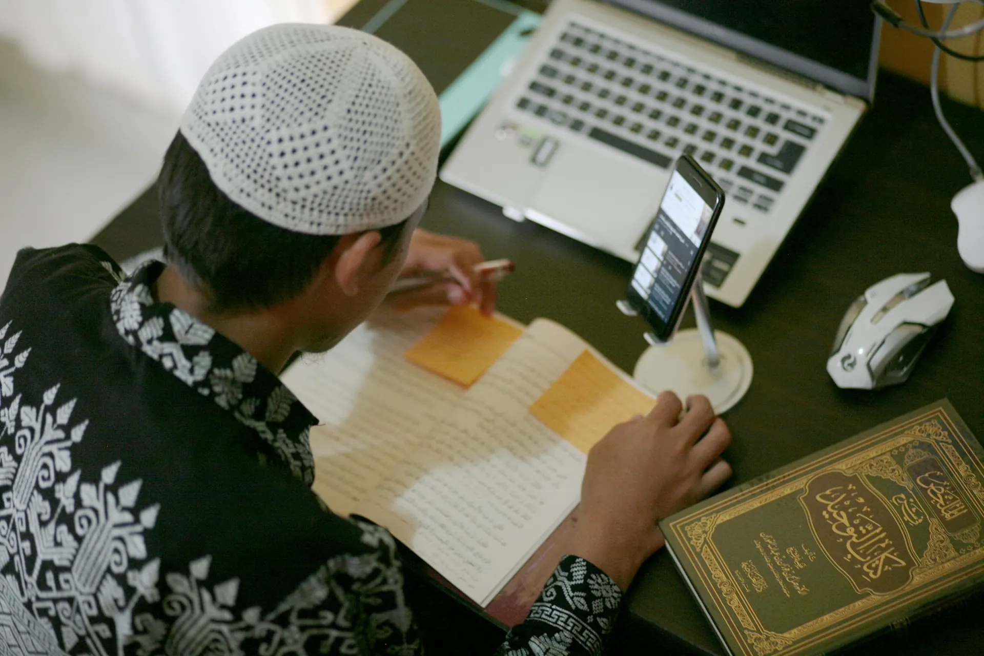 طالب يدوّن ملاحظات في دفتر بجانب حاسوب محمول وكتاب